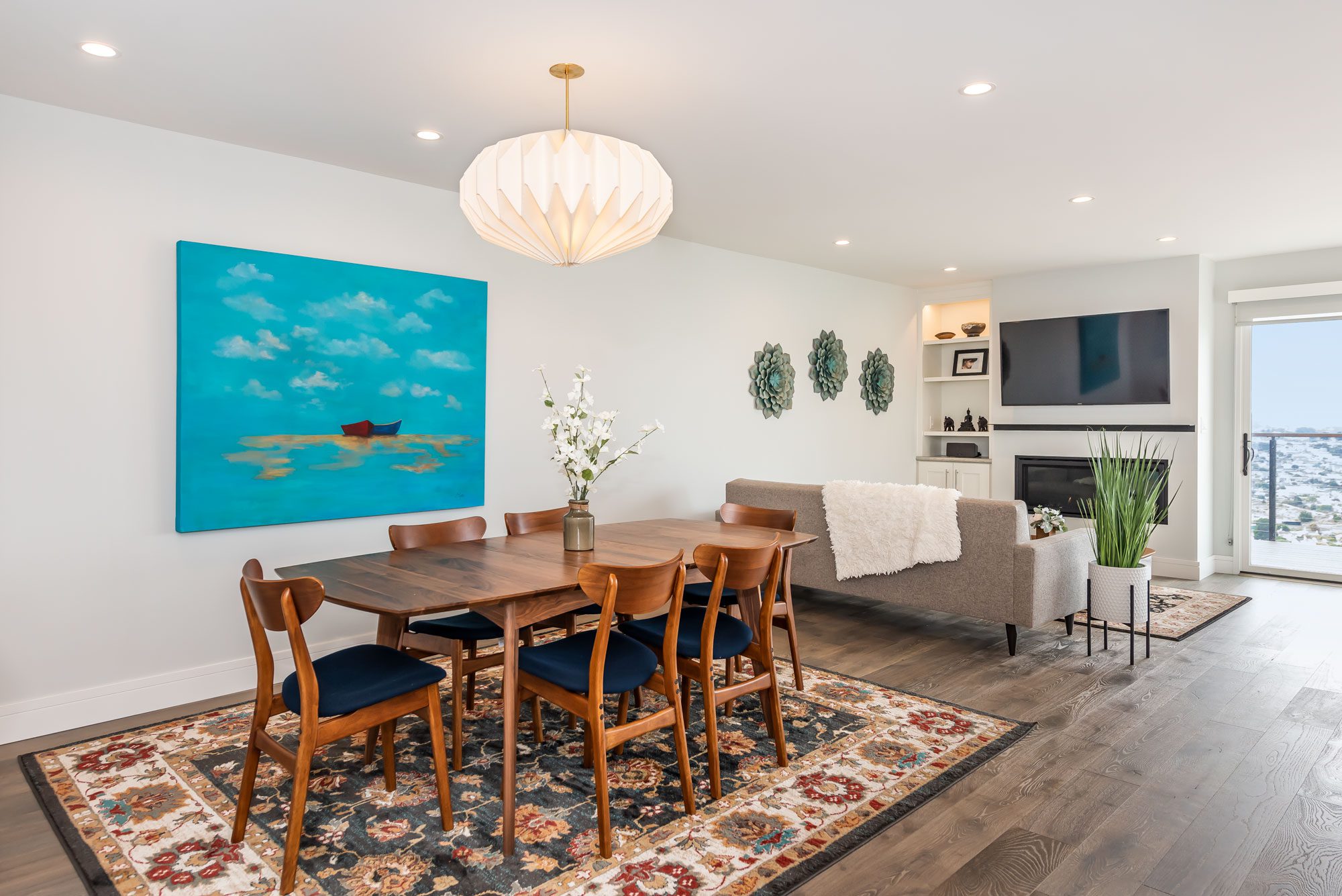 A bright, modern dining room with a wooden table and chairs, a large blue artwork on the wall, and a view of the city outside.