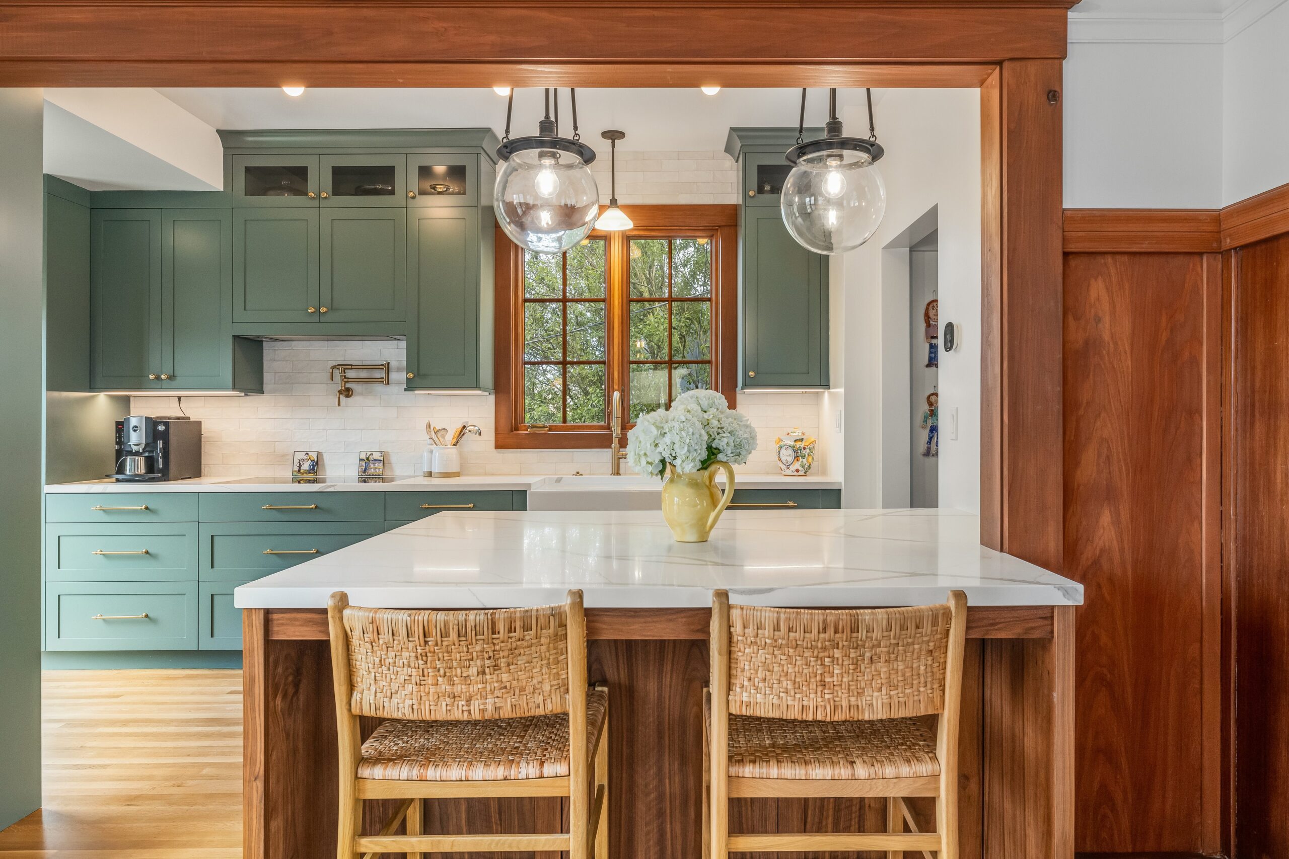 Modern kitchen with teal cabinetry, white countertops, rattan bar stools, and pendant lights, viewed from an adjacent room with wooden trim.