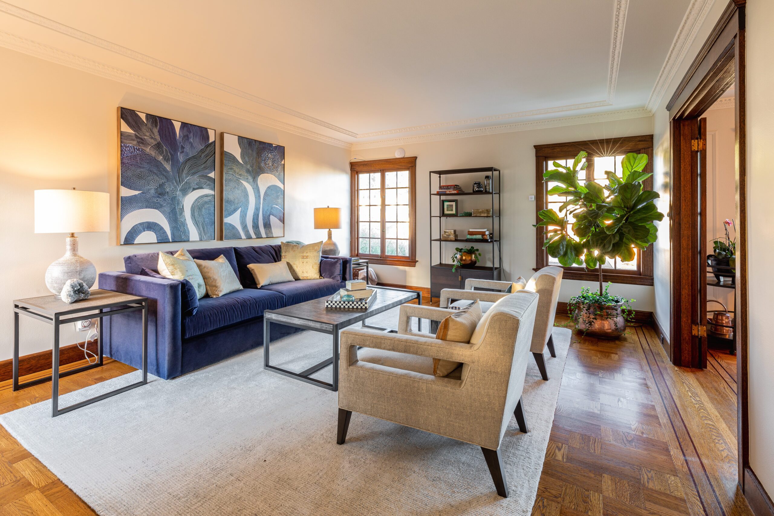 Elegant living room with a blue sofa, two beige armchairs, a large wall painting, and several plants, featuring hardwood floors and natural lighting.