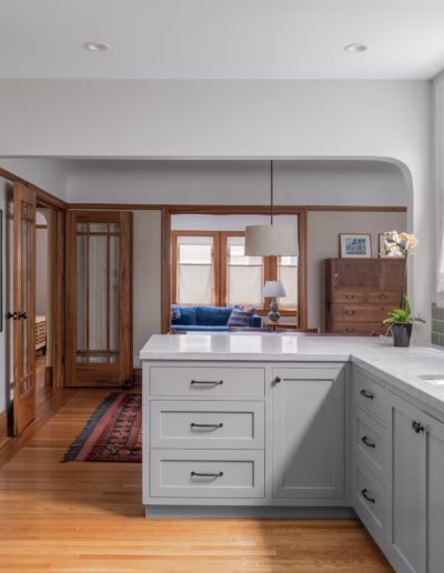 Modern kitchen with white cabinets, green tile backsplash, black faucet, and large windows; view extends to a living area with wood trim and furniture.