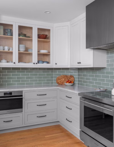 Modern kitchen with light gray cabinets, green subway tile backsplash, stainless steel appliances, wooden floor, and a loaf of bread on the counter.