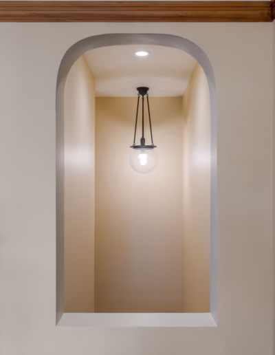 A glass pendant light hangs from the ceiling in a recessed wall niche with an arched opening and beige walls.