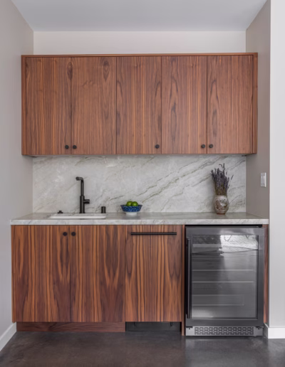 Modern kitchenette with wood cabinets, marble backsplash, black faucet, small sink, glass-front mini fridge, and a vase with dried flowers on the counter.
