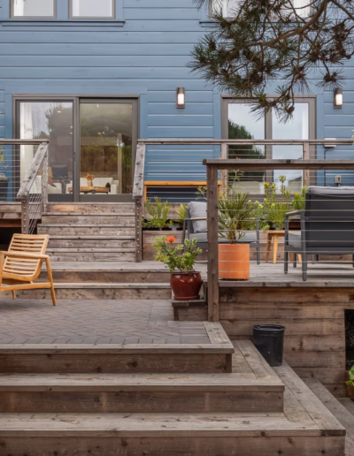 Wooden multi-level backyard deck with outdoor seating areas, potted plants, and stairs leading up to a blue house with large windows and glass doors.