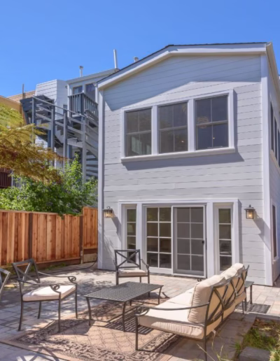 A two-story gray house with a fenced backyard patio, outdoor seating, a grill, and a small tree, under a clear blue sky.