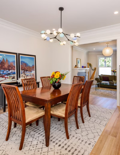 A dining room with a wooden table and six chairs, a modern chandelier, two framed pictures on the wall, and a cabinet with a fishbowl on top.