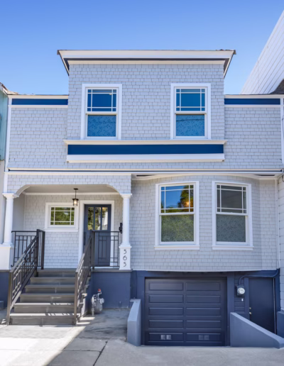Two-story light gray house with white trim, blue accents, front steps leading to the entrance, and a dark garage door on the ground level.