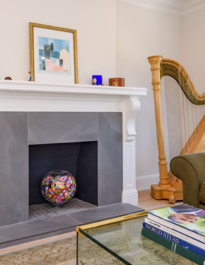 A living room with a gray fireplace, a glass bowl filled with colorful items, a harp near a window, a green sofa, and a glass coffee table with books.