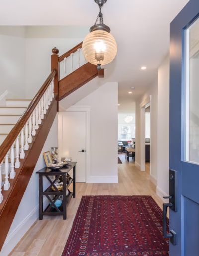 A front entryway with a blue door, wooden staircase with white spindles, red patterned rug, and a small table displaying framed photos and decor.