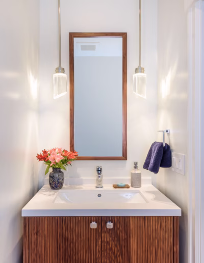 A small bathroom vanity with a wood cabinet, white sink, rectangular mirror, two pendant lights, a vase of flowers, soap dispenser, and a navy towel on the wall.