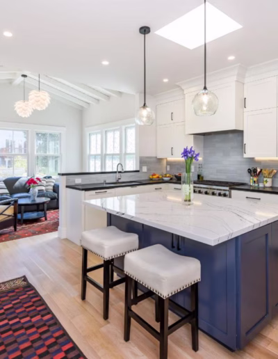 Modern kitchen with white cabinets, a blue island with two stools, pendant lights, and an open living area with a sofa and large windows in the background.