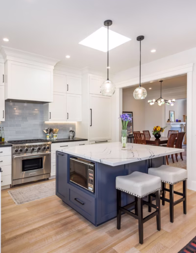Modern kitchen with white cabinets, stainless steel appliances, marble island with barstools, hardwood floors, pendant lights, and a view into a dining room.