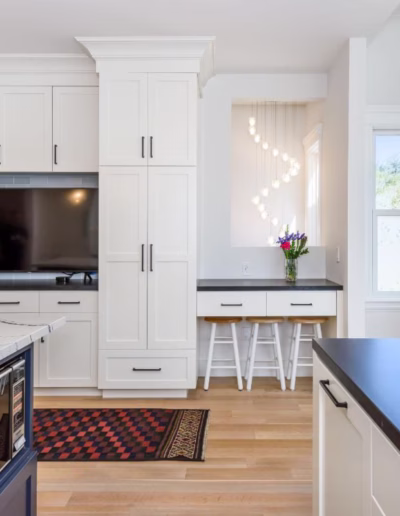 Modern kitchen with white cabinets, black countertops, a built-in TV, a vase of flowers, and natural light from large windows.