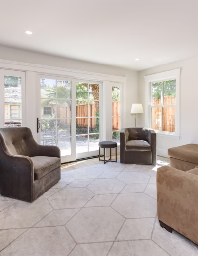 A bright living room with a beige sectional sofa, two dark armchairs, a round ottoman, a TV, and large windows overlooking a fenced backyard.