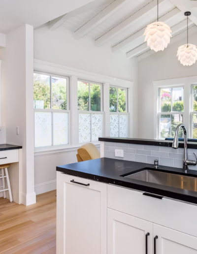 Modern kitchen with white cabinets, black countertops, a stainless steel sink, pendant lights, and a small desk area with two stools by the windows.