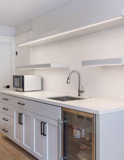 Modern kitchen with white cabinets, built-in sink, microwave, glass-front mini fridge, under-shelf lighting, and white subway tile backsplash. Light wood flooring.